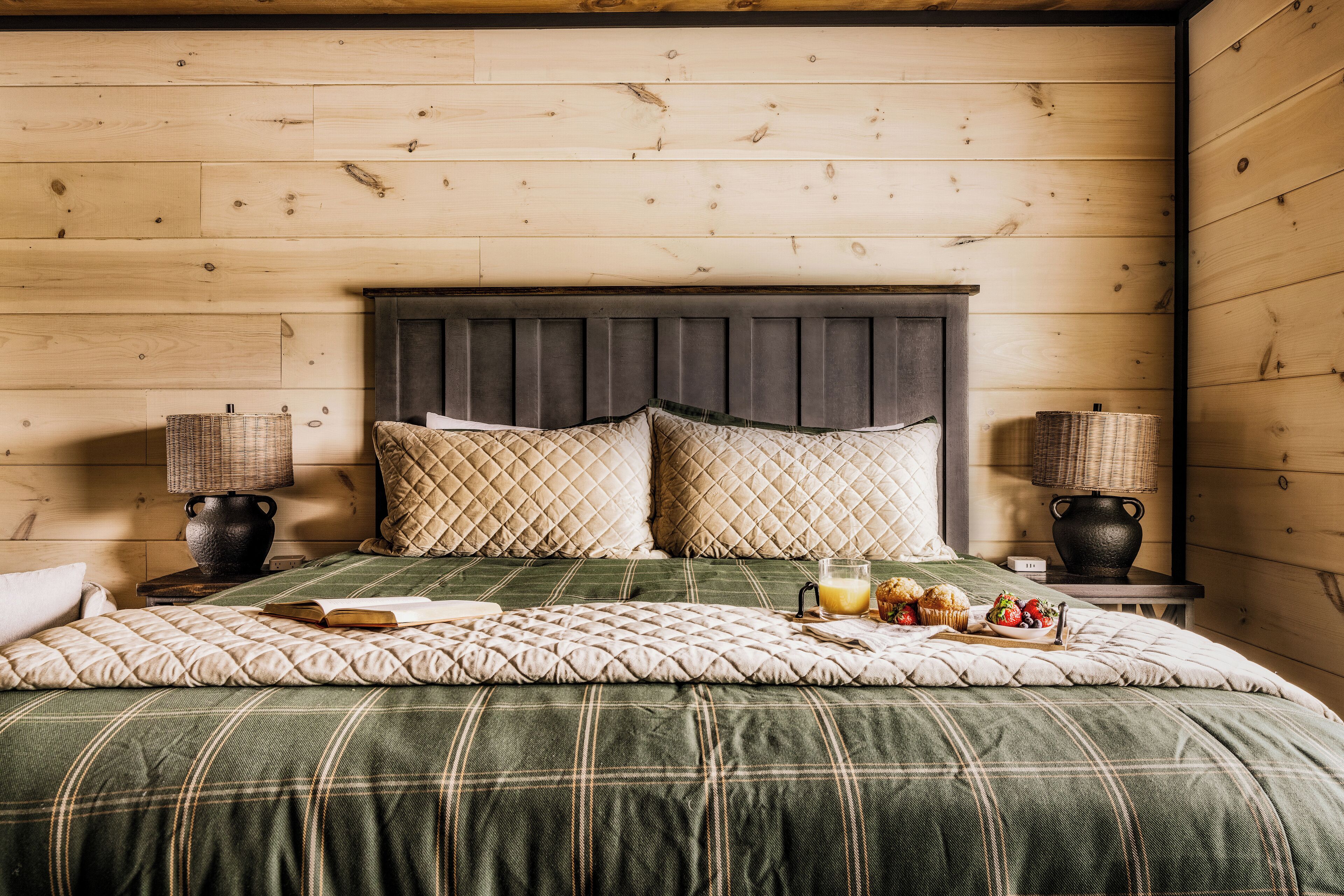 A neatly made bed with a plaid bedspread, two pillows, a bedside lamp, and a basket of muffins on a wooden wall.