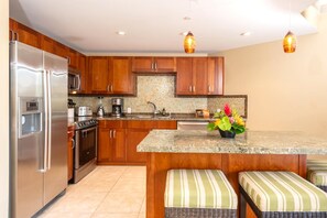 A modern kitchen with wooden cabinets, stainless steel appliances, a granite countertop, and a central island with a woven bench. - Honua Kai | Ocean View 5 BR, Sleeps 16 | Car Incl. w/ 6+ Nights | HKH ML-1784 by KBM (Lahaina)