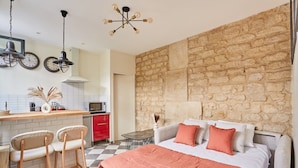 A bedroom with a stone wall, a bed with orange bedding, and a kitchen area with a red cabinet and a clock. - Sublim Apartement Center of Paris (Paris)