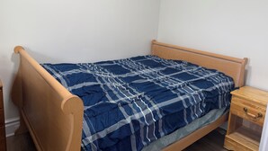 En säng för en person med en träram och ett blått, mönstrat överkast i ett rum med vita väggar och ett träbord vid sängen med en låda.