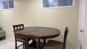 A dining area with a round table and four chairs, beige walls, and two windows with blinds.