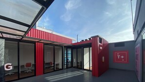 Un edificio moderno con fachada roja, puertas de cristal y un letrero T+ que indica zona para fumadores.