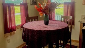 Un coin repas avec une table ronde, des chaises, un vase de fleurs et une vue sur un paysage verdoyant par les fenêtres.