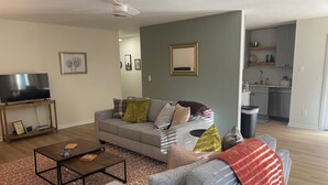 A living room with a grey sofa, a coffee table, and a TV on a stand. There is a kitchen in the background.