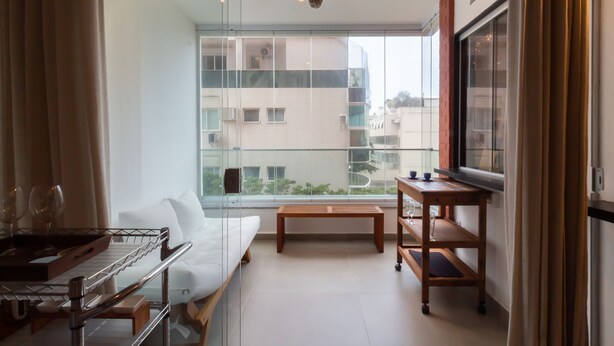 A modern hotel room with a large window, a bed, a wooden bench, a small table with glasses, and a shelf with bottles.