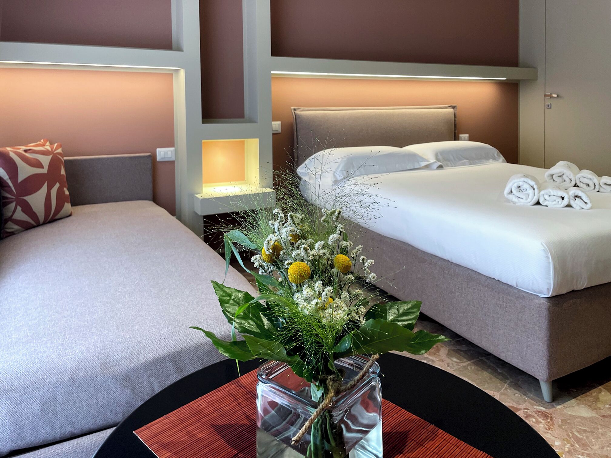 A hotel room with two beds, a bedside table with a vase of flowers, and a neatly made bed with towels.