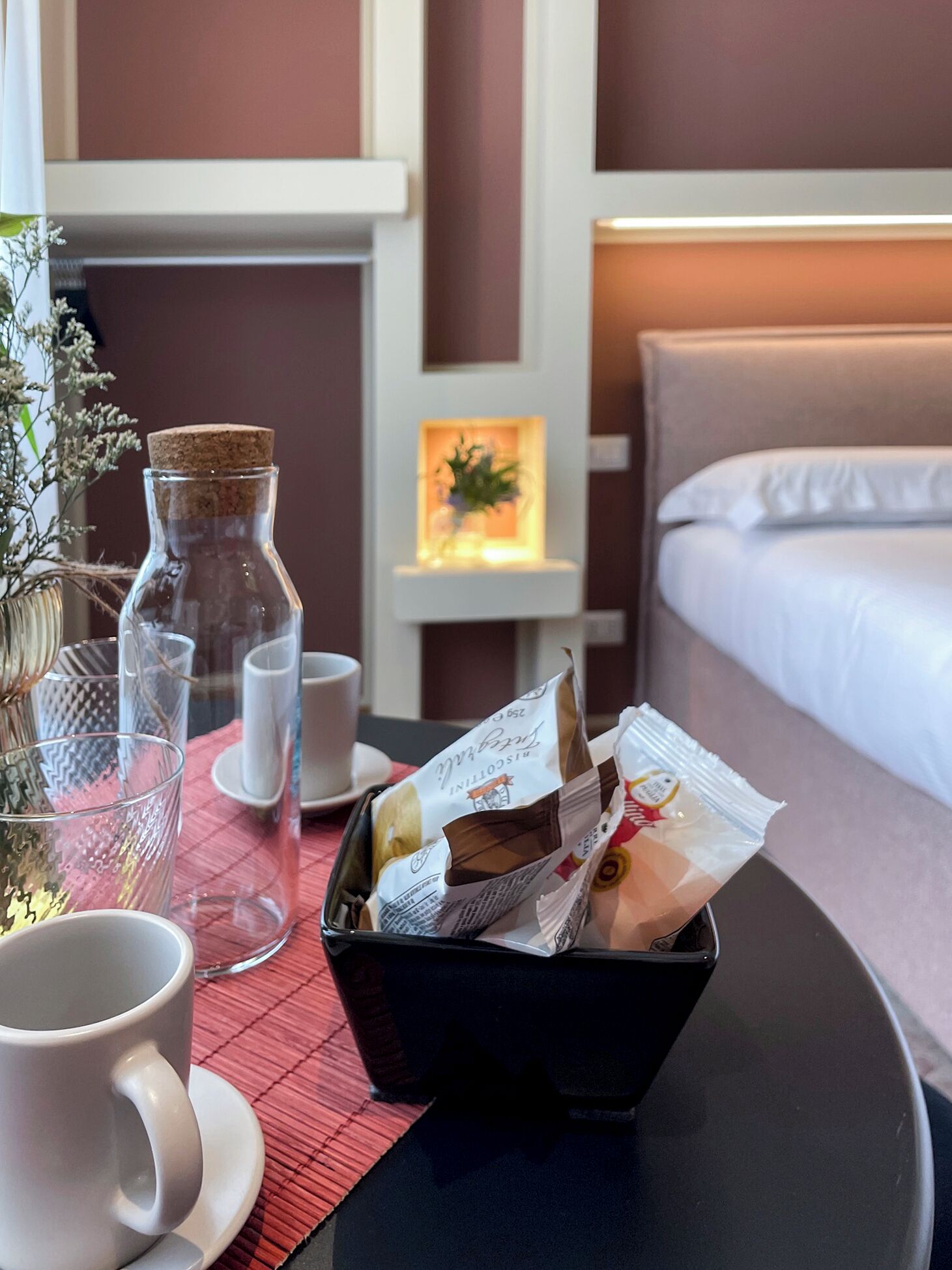 A hotel room with a bed, a bedside table with a lamp, a vase with flowers, a coffee cup, and a basket of breakfast items.