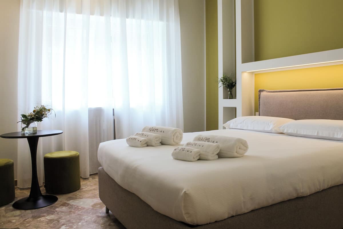 A neatly made bed with white linens and towels, a small round table with a vase of flowers, and a green ottoman.