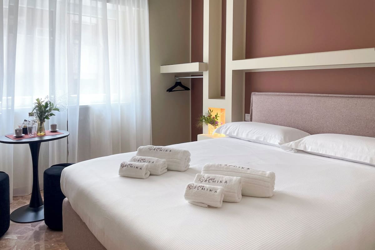 A hotel room with a neatly made bed, a small table with a vase and cups, and a hanging coat rack.