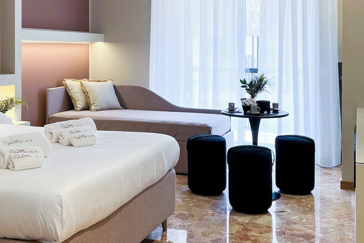 A modern hotel room with a large bed, a sofa, a small table with a plant, and a view of the city through sheer curtains.