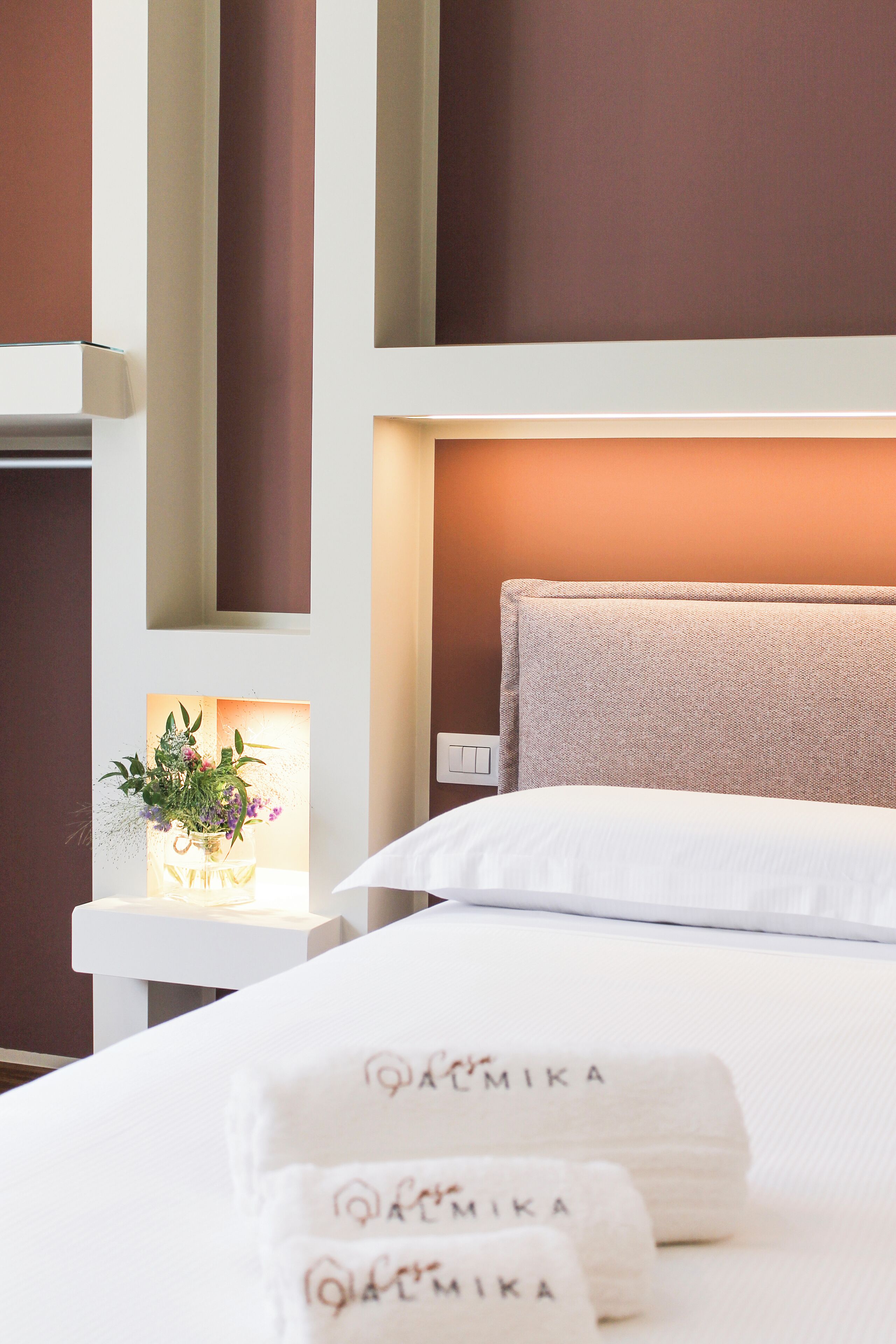 Cama bem arrumada com roupas de cama e toalhas brancas, mesa de cabeceira com um vaso de flores e uma luminária na parede.