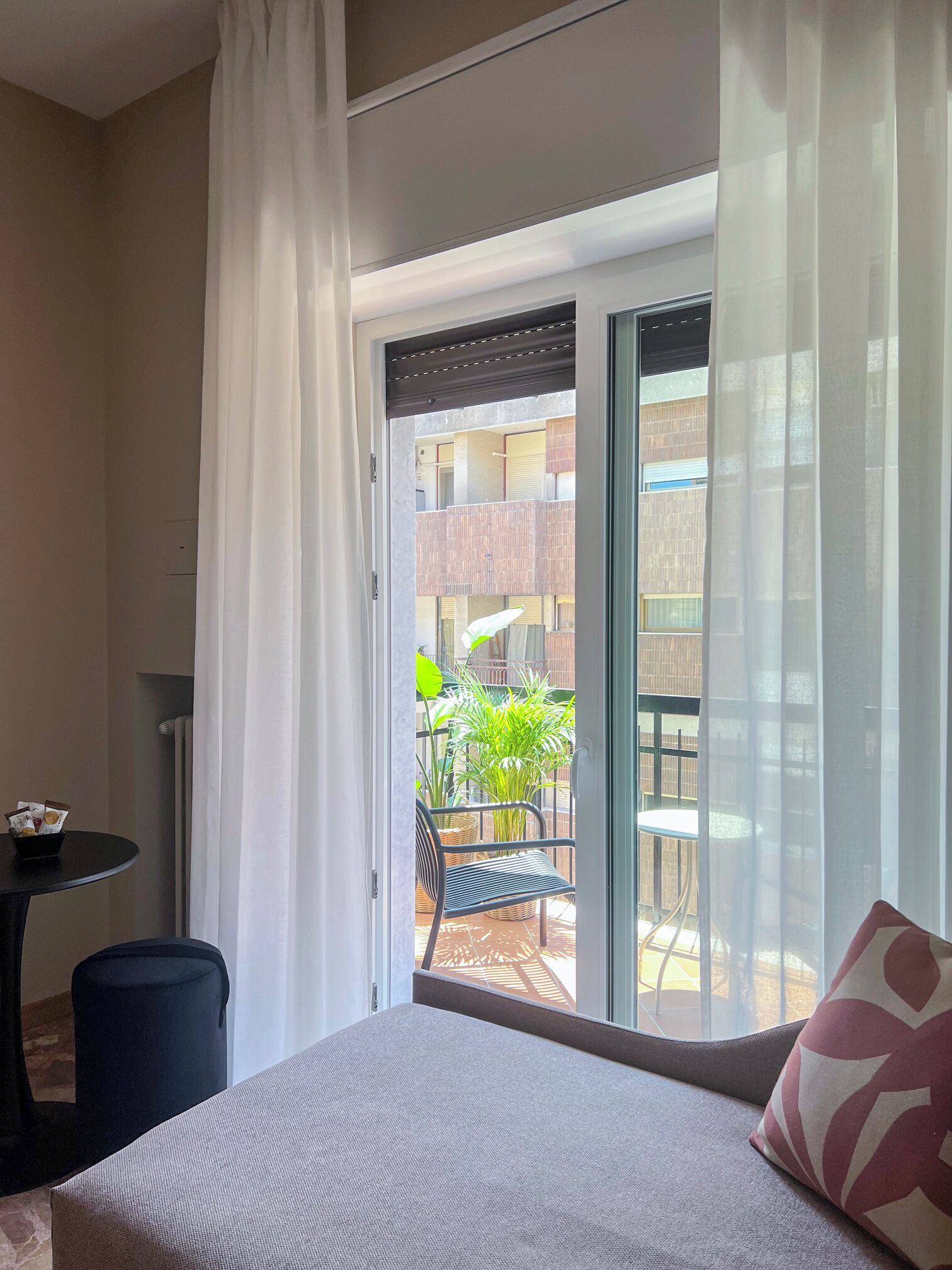 Une chambre d’hôtel avec un lit, une petite table sur laquelle se trouve un panier rempli de divers objets, une chaise, et une vue sur un balcon extérieur agrémenté de plantes et sur lequel on aperçoit un bâtiment au loin.