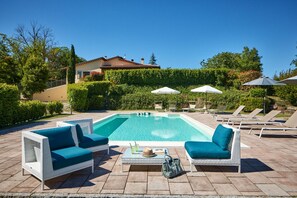 A swimming pool with lounge chairs and umbrellas, surrounded by greenery and a building in the background.