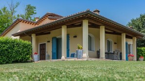 A two-story house with a tiled roof, a covered porch with chairs, and a well-maintained lawn.