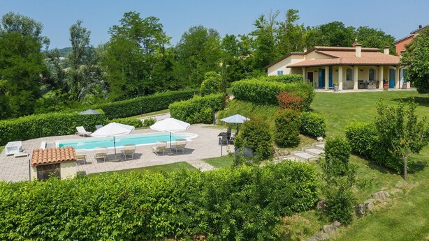 A pool area with lounge chairs and umbrellas, surrounded by well-maintained hedges and a building with a tiled roof.