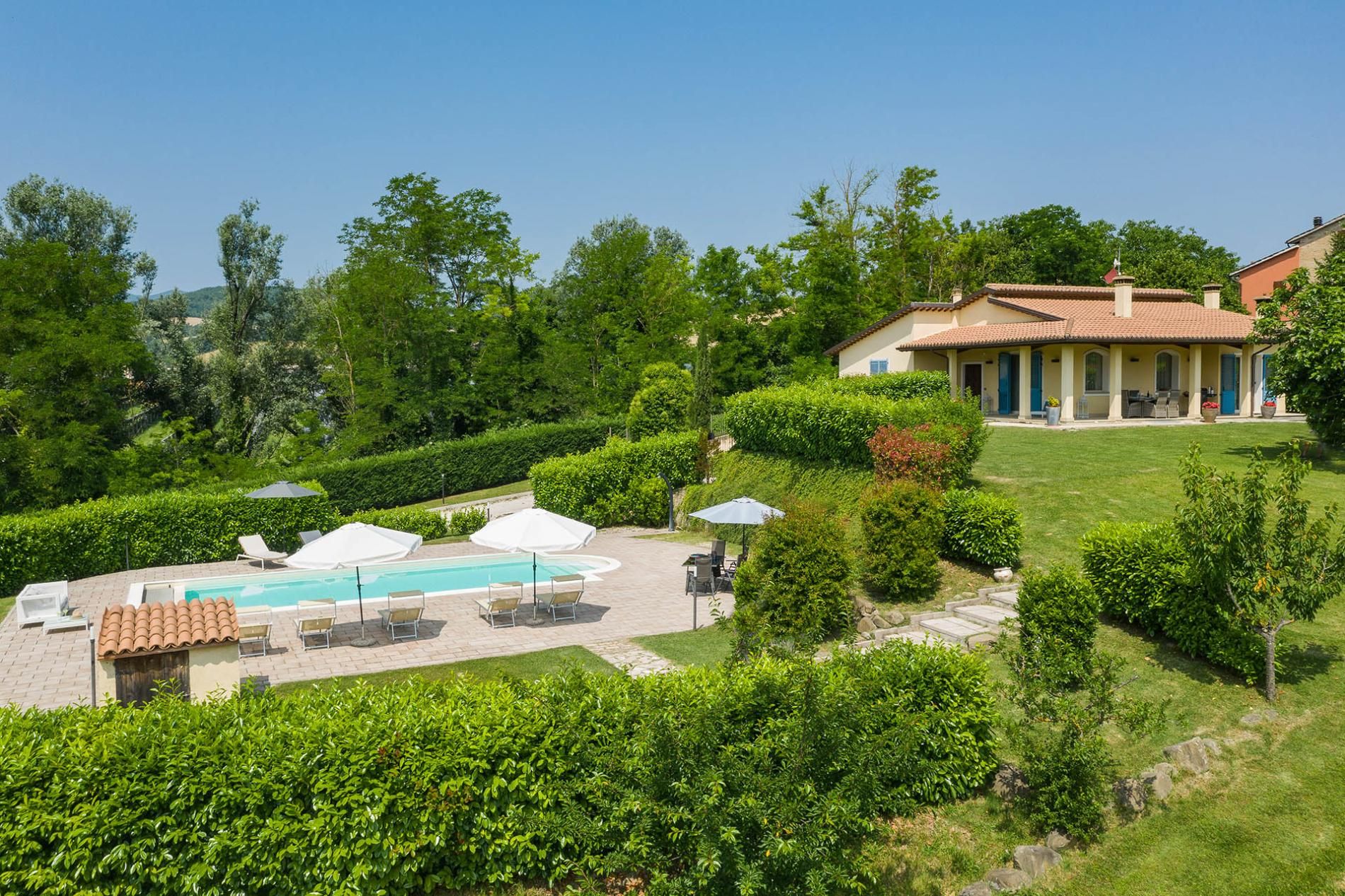 A pool area with lounge chairs and umbrellas, surrounded by well-maintained hedges and a building with a tiled roof.