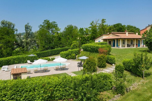 A pool area with lounge chairs and umbrellas, surrounded by well-maintained hedges and a building with a tiled roof.