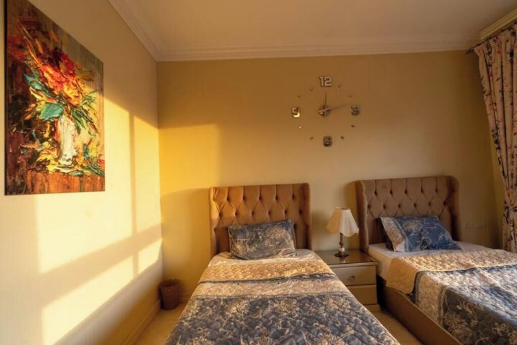 Habitación de hotel con dos camas, un cuadro grande en la pared, un reloj y una ventana con cortinas.