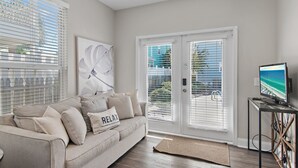 A living room with a beige sofa, a large window with blinds, a flat-screen TV on a stand, and a view of the outdoors.
