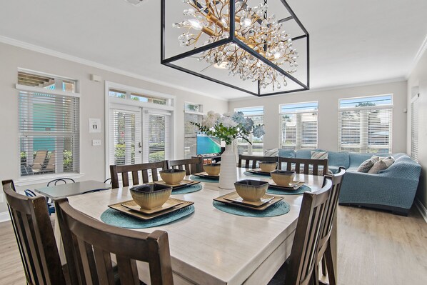 A dining area with a round table, chairs, and a chandelier. There is a sofa and a window with blinds in the background.
