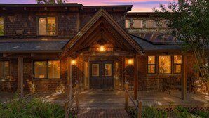 A rustic wooden house with a prominent front porch, large windows, and a brick pathway leading to the entrance.
