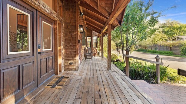 A wooden porch with a bench, a welcome mat, and a view of a road and greenery.