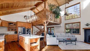 A spacious living area with a large white sofa, a coffee table, and a fireplace. There is a kitchen with wooden cabinets and a central island. The room has large windows and a wooden ceiling.