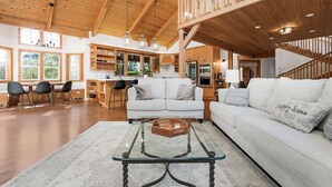 A spacious living area with a high ceiling, wooden beams, a central seating area with a glass coffee table, and a kitchen in the background.