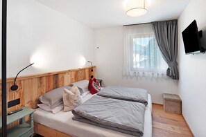 A bedroom with a wooden headboard, two beds, a bedside table with a lamp, a TV mounted on the wall, and a window with curtains.