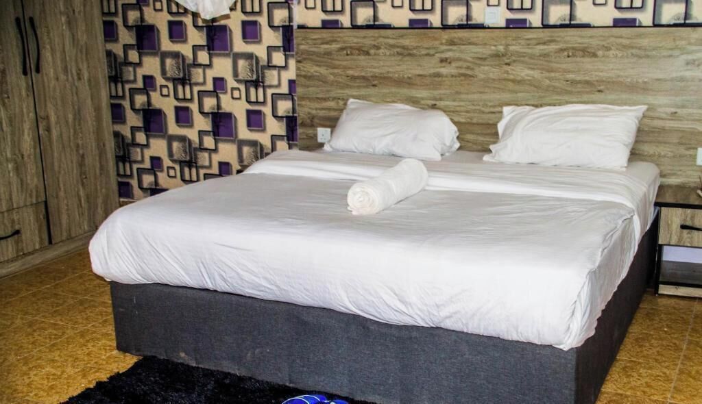 A neatly made bed with white linens and a rolled towel, a pair of blue slippers on a dark rug, and a patterned wall in the background.