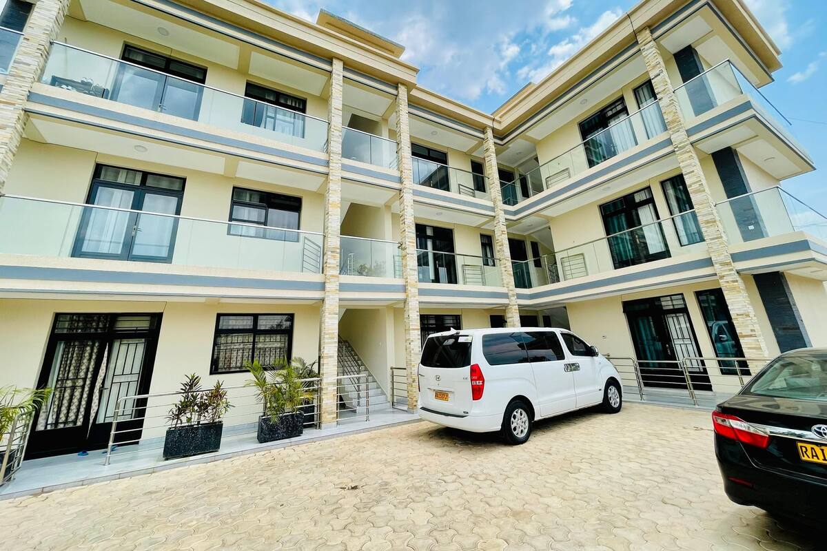 A multi-story residential building with balconies, a car parked in front, and potted plants.