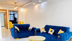 Een moderne woonkamer met een blauwe zithoek, een ronde salontafel met gouden lijst en een grijze fauteuil.