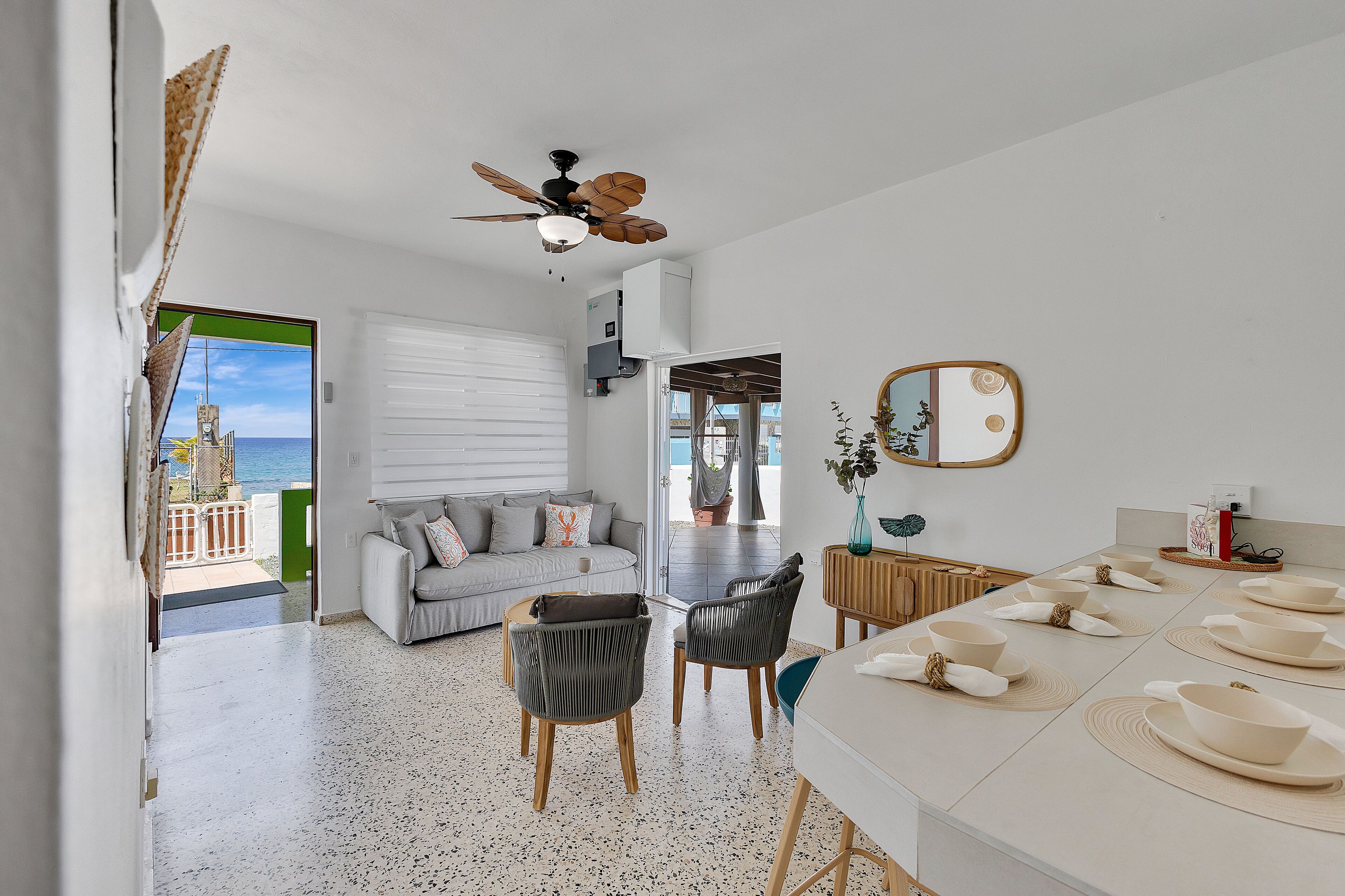 A modern living room with a sofa, chairs, a dining table with bowls and napkins, a ceiling fan, and a view of the sea through a door.