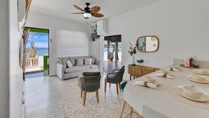 A modern living room with a sofa, chairs, a dining table with bowls and napkins, a ceiling fan, and a view of the sea through a door. - Sunset Marena/Beach Front House (Stella)