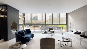 Una sala de estar moderna con un sofá, sillones y mesas de centro. Amplias ventanas ofrecen vistas al exterior.