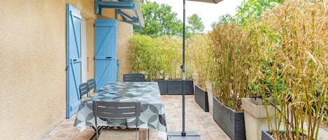 Een terras buiten met een tafel, stoelen, een grote parasol en een muur met blauwe luiken.