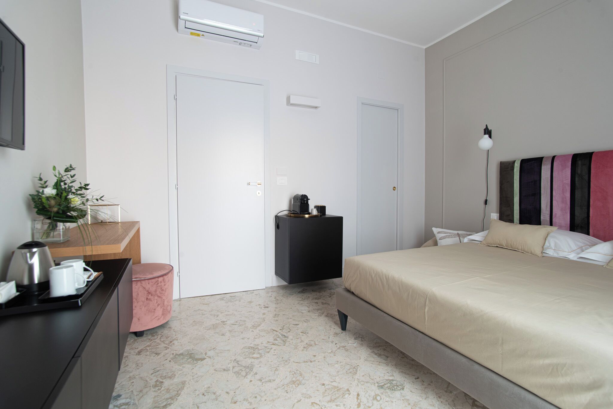 Un dormitorio con cama, mesita de noche y un escritorio pequeño con silla. Se observa una puerta y un equipo de aire acondicionado en la pared.