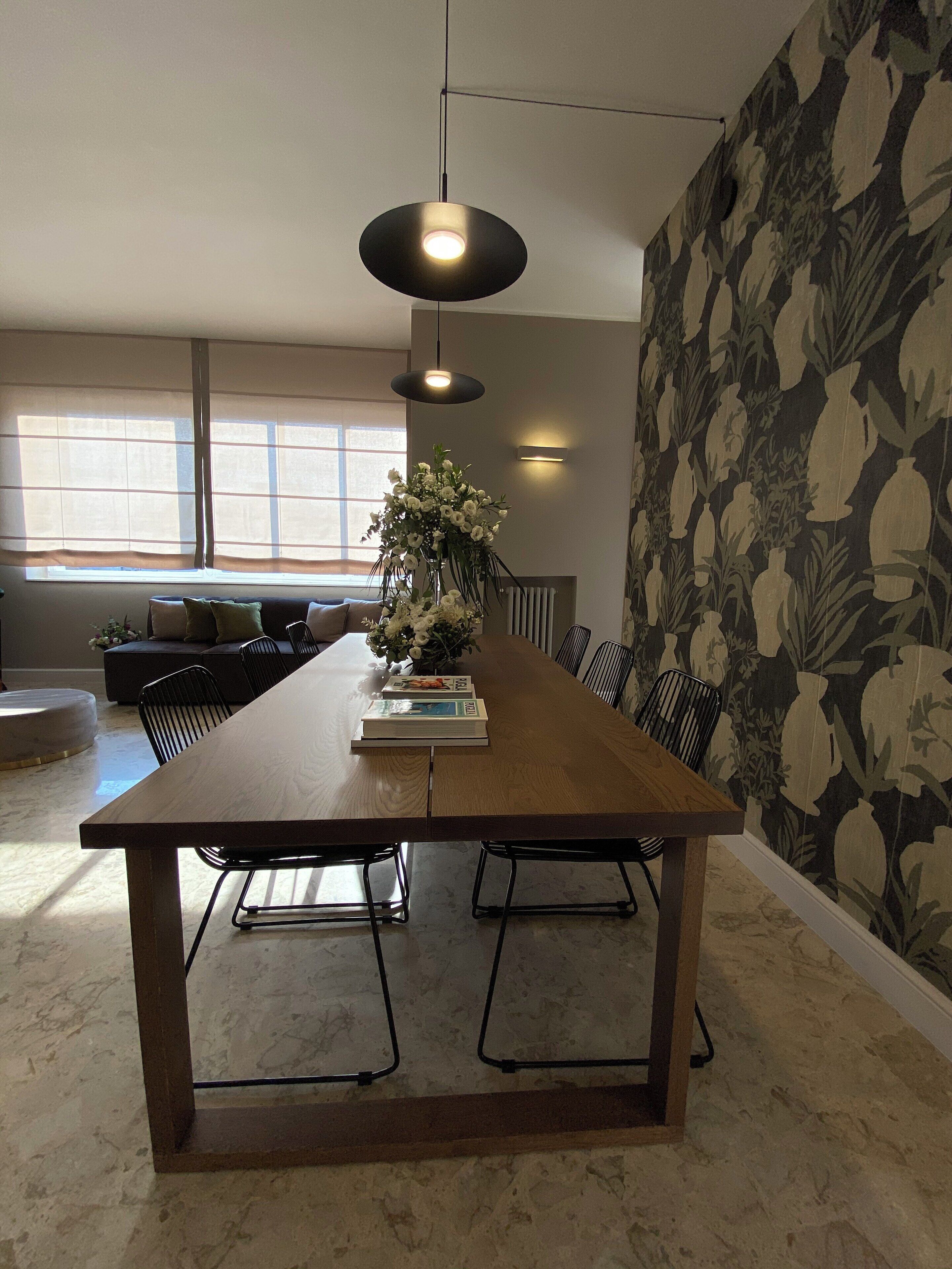 Un comedor con una larga mesa de madera, sillas negras, una lámpara colgante y un arreglo floral.