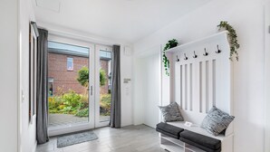 Una habitación moderna con un ventanal corredera que da a un jardín, un banco blanco con cojines y un estante blanco montado en la pared con ganchos.
