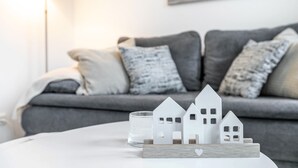 Un salón moderno con un sofá gris, cojines blancos y un pequeño modelo de casa blanca sobre una mesa blanca.