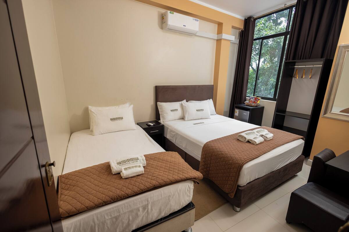 A hotel room with two beds, each with a brown quilted bedspread and white linens. There is an air conditioning unit on the wall, a small table with a lamp, and a window with curtains.
