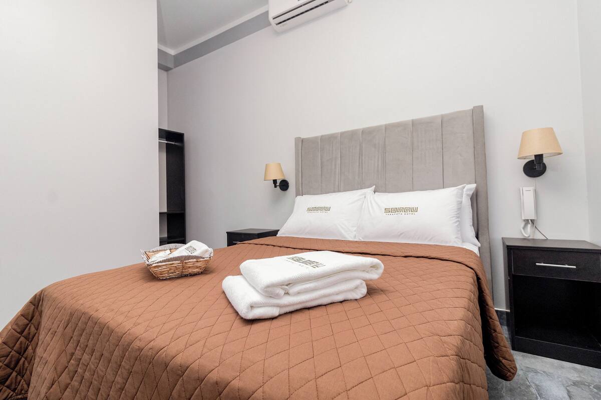 A neatly made bed with a brown quilt, white pillows, and folded towels on a headboard. There are two bedside tables, one with a lamp and the other with a phone.