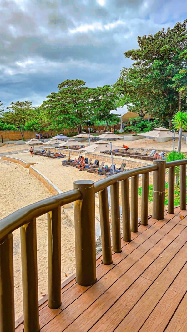 Una terraza frente a la playa con sillones y sombrillas, rodeada de exuberante vegetación y una zona de arena.