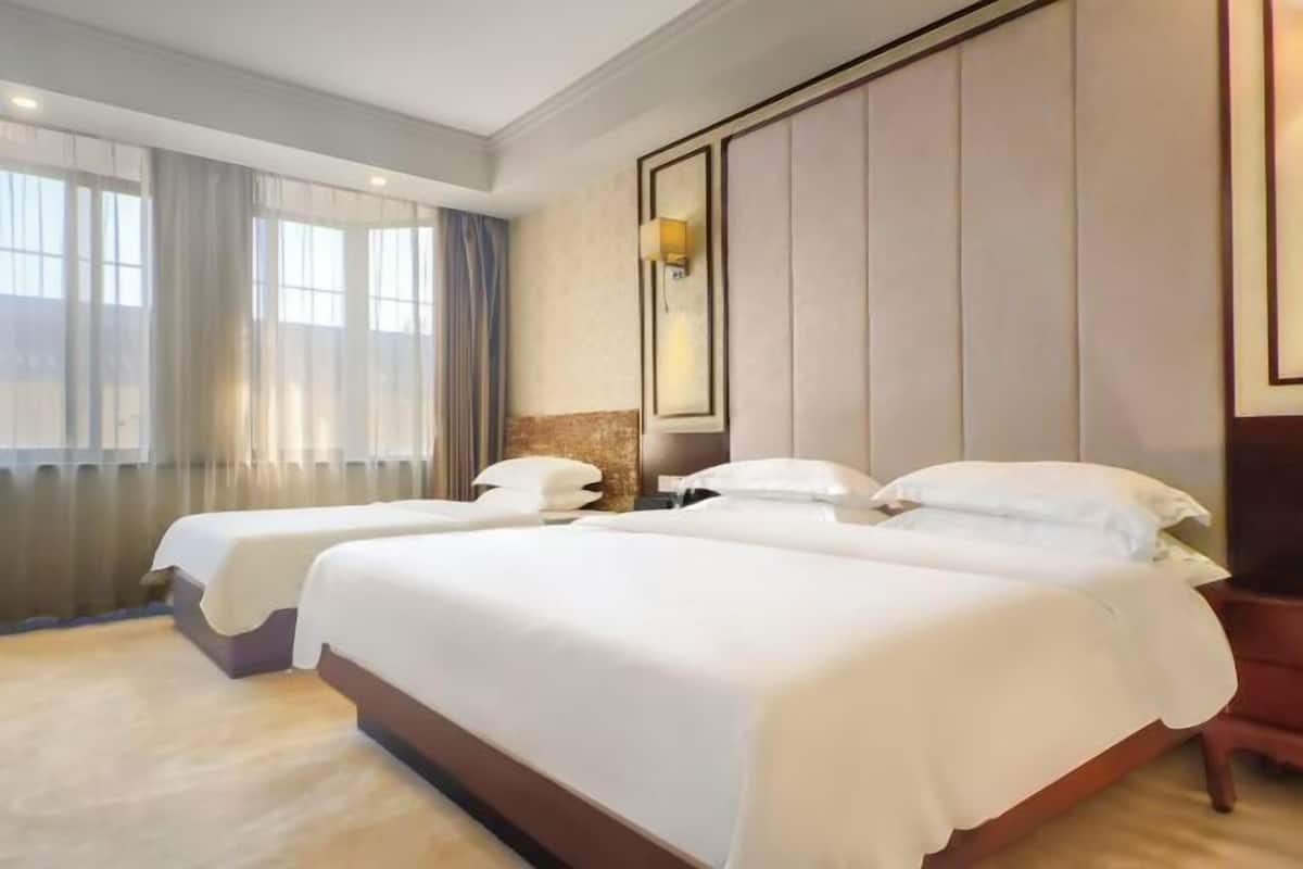 A hotel room with two beds, a large window with curtains, a wall-mounted light, and a wooden nightstand.