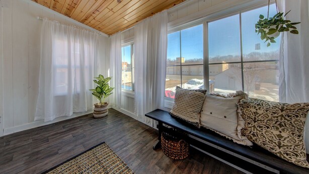 A cozy room with a wooden ceiling, large windows with white curtains, a bench with cushions, and a potted plant. - Charming Downtown Retreat for Weddings, Weekends & Outdoor Fun! (Delano)
