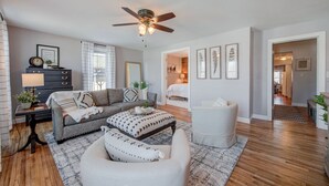A living room with a sofa, ottoman, and a ceiling fan. There is a dresser with a lamp and a plant. The room has wooden flooring and a rug. - Charming Downtown Retreat for Weddings, Weekends & Outdoor Fun! (Delano)