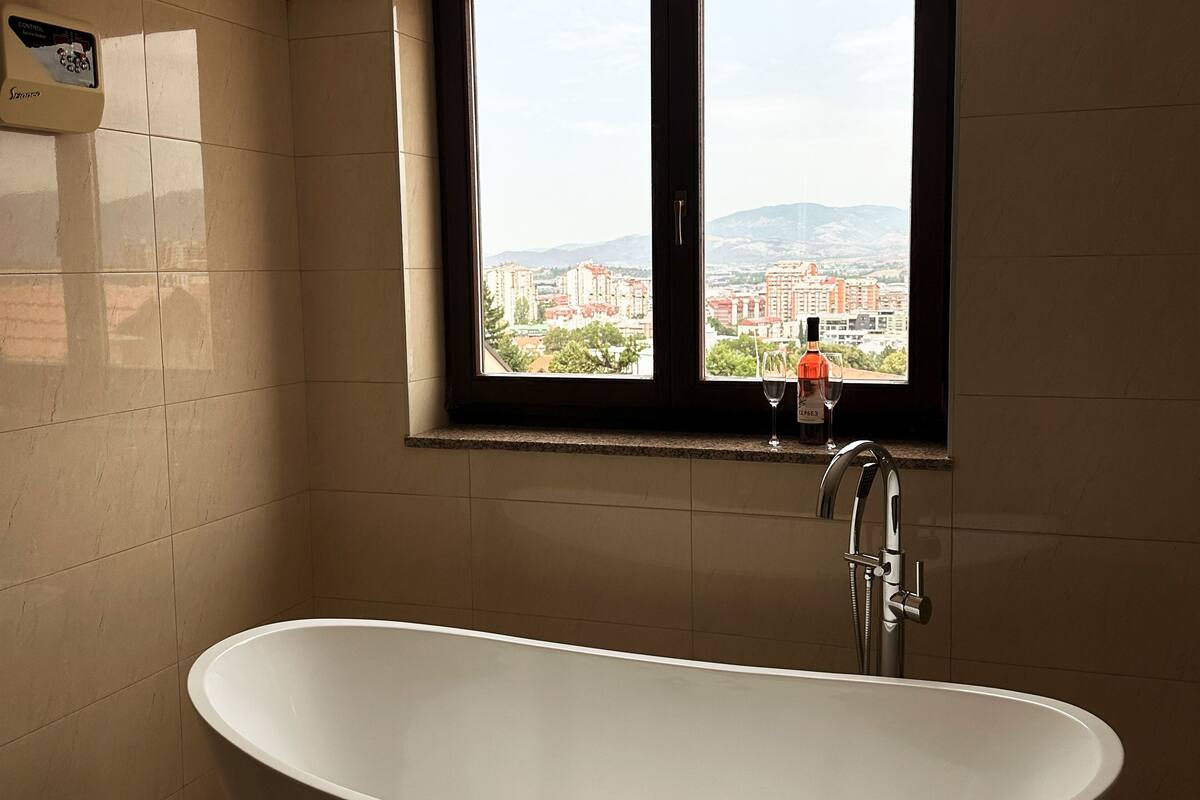 Ein Badezimmer mit freistehender Badewanne, einem Fenster mit Blick auf Gebäude und einer Bedienkonsole an der Wand.