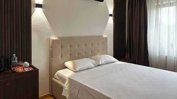 A hotel room with a large bed, a bedside table with a water bottle, glasses, and snacks, and a ceiling-mounted air conditioner. - Hillside Garden (Skopje)