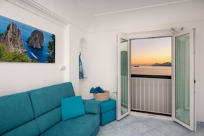 A balcony with a view of the sea and sunset, a teal sofa, and a painting of a coastal landscape.
