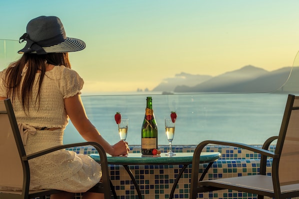 A person seated at a table with a bottle of champagne and glasses, overlooking a body of water with mountains in the distance.